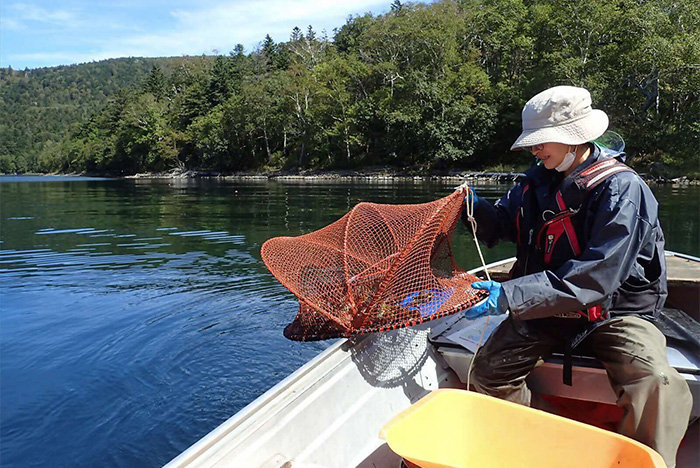 然別湖生物多様性保全協議会の活動を写した写真。湖に浮かぶボートに、アウトドアウェアを着た人が1人写っている。捕獲用の網を手に持ち、中を覗き込んでいる。