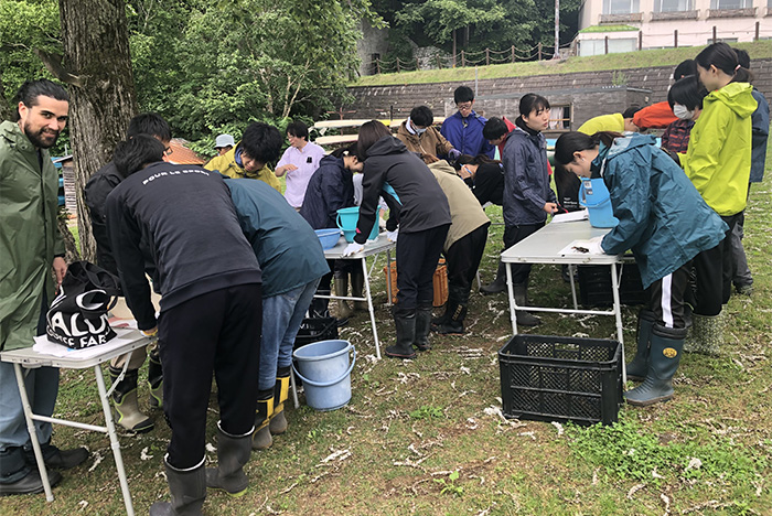 然別湖生物多様性保全協議会の活動を写した写真。アウトドアウェアを着た沢山の人が、キャンプ用折り畳みテーブルの上に載ったバケツや、ウチダザリガニを覗き込んでいる。